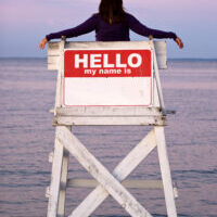 A woman relaxes in a vacant lifeguard chair where the sign on the back reads HELLO MY NAME IS.